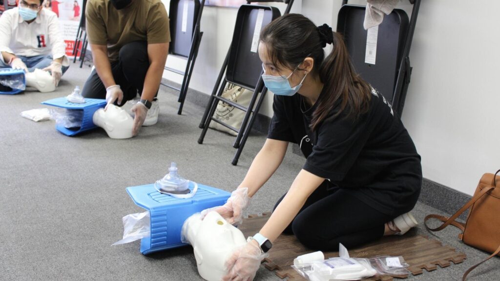 People wearing masks kneel on the floor during a CPR training session. They practice chest compressions on CPR mannequins placed on mats. Chairs are visible in the background.