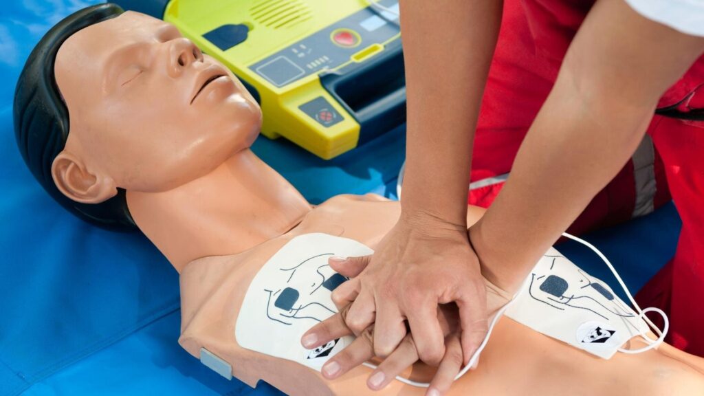 A person is demonstrating CPR on a training mannequin. They are pressing down on the chest with both hands. An AED device is visible in the background on a blue mat. The mannequin has pads placed on its chest.