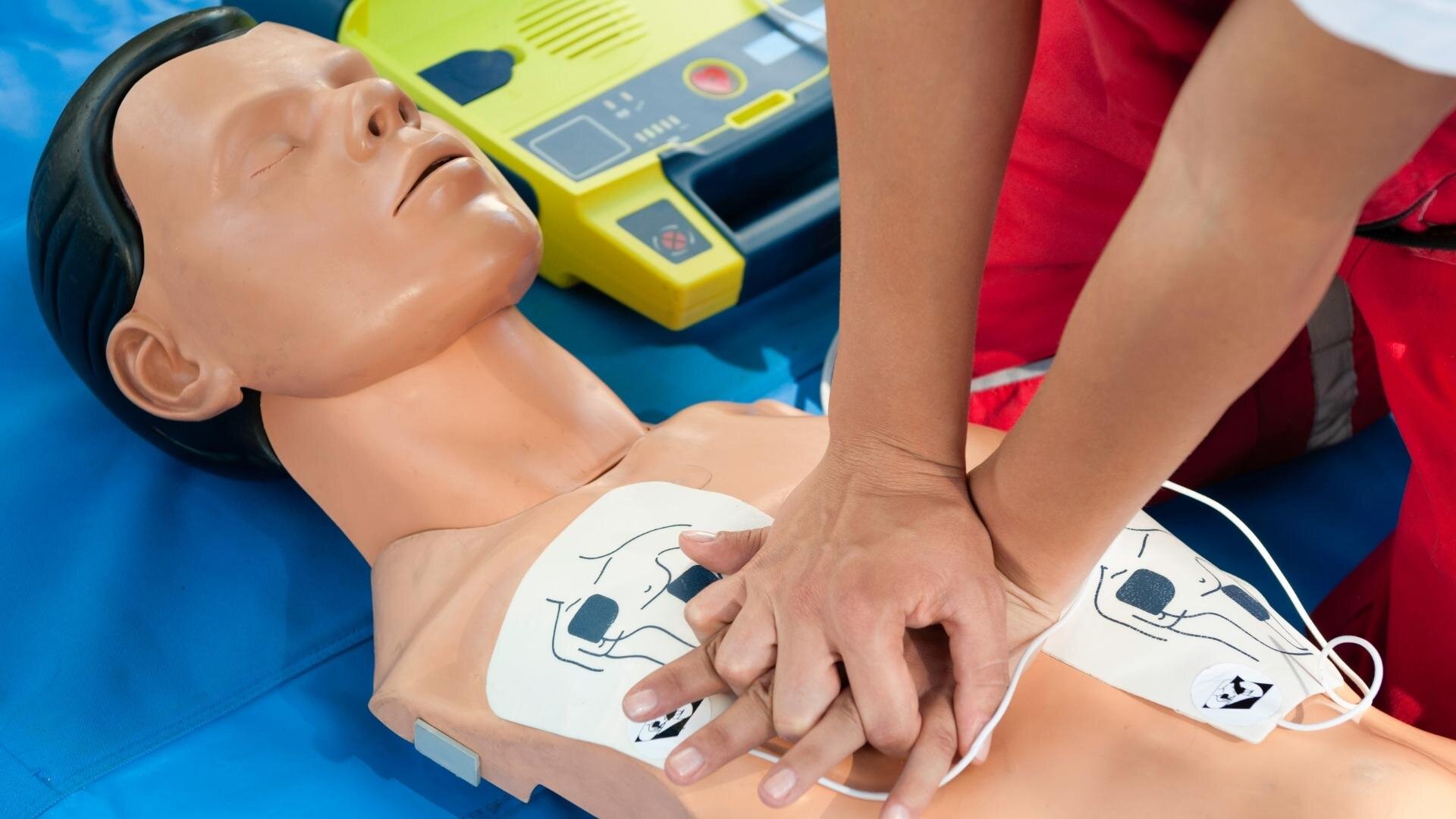A person is demonstrating CPR on a training mannequin. They are pressing down on the chest with both hands. An AED device is visible in the background on a blue mat. The mannequin has pads placed on its chest.