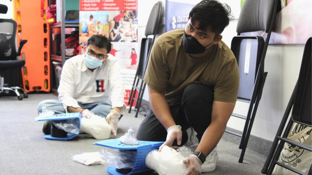 Two individuals wearing masks and gloves practice CPR techniques on dummies placed on the floor. They are kneeling, each with a dummy in front of them. Posters and medical equipment are visible in the background.
