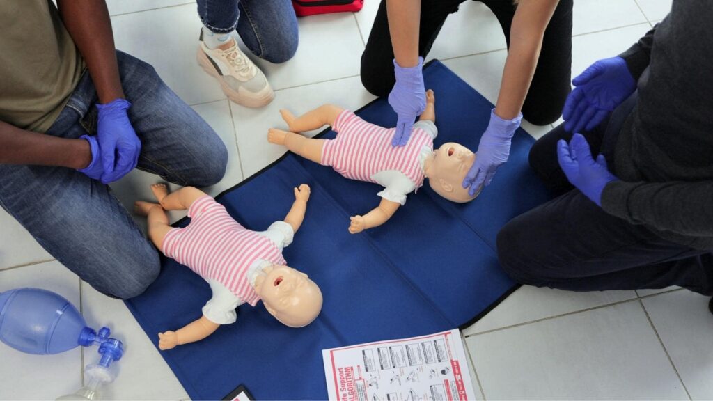 People are kneeling on the floor, wearing blue gloves, practicing CPR on two baby mannequins dressed in striped outfits. A set of instructions and a medical bag are nearby.