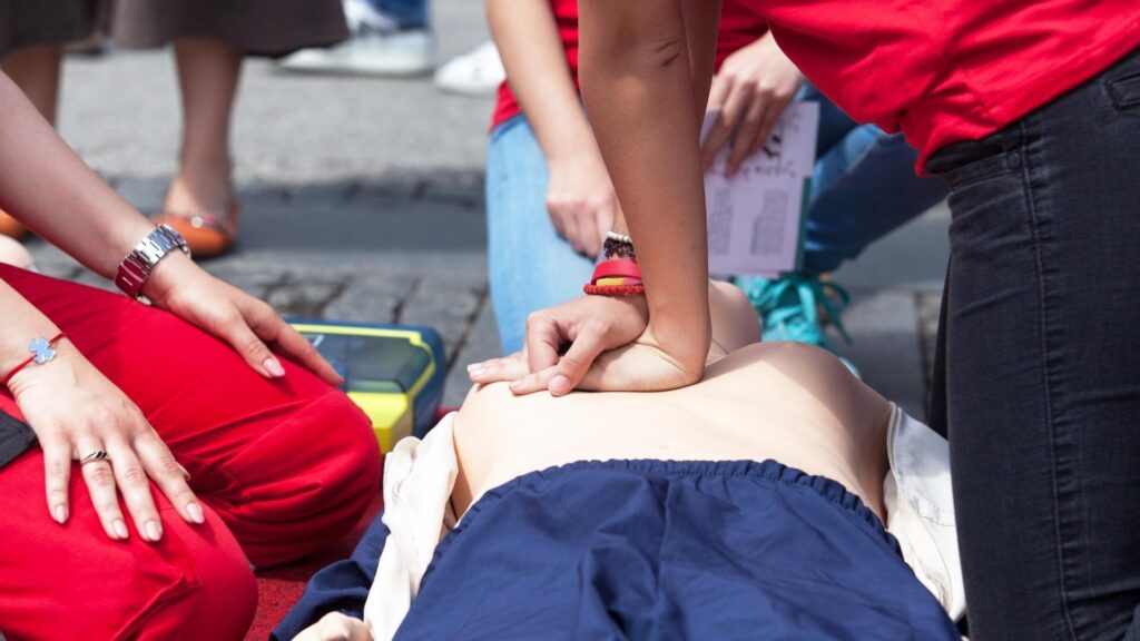 A person practices CPR on a mannequin outdoors. They press on the chest, surrounded by others observing. Nearby, a defibrillator is visible. People wear red shirts, suggesting a training or demonstration event.