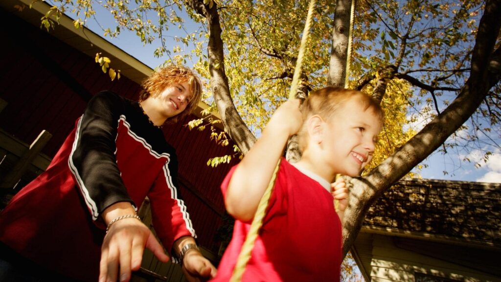 A teenager pushes a young boy on a tree swing. Both are smiling and wearing red shirts. The swing is in a backyard with a tree and house in the background under a clear blue sky. The scene is sunlit and cheerful.