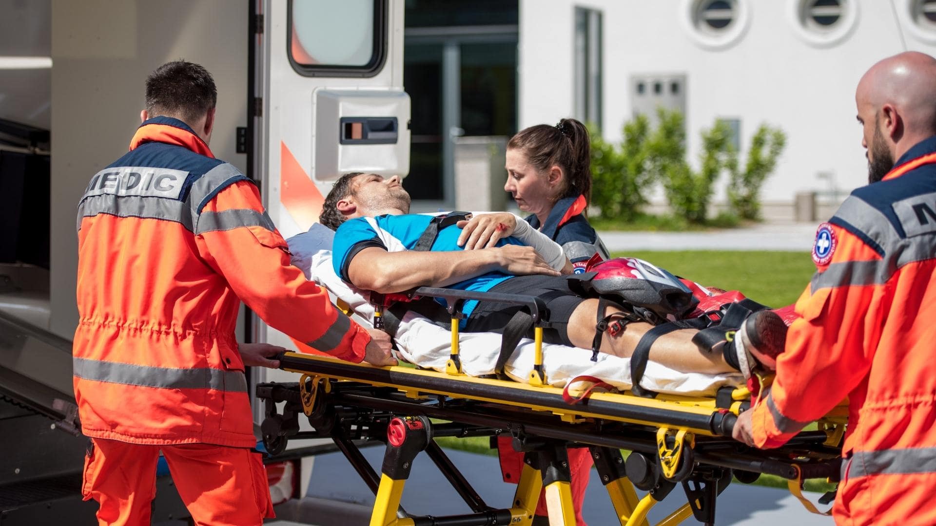 Three paramedics in bright orange uniforms transport an injured cyclist on a stretcher into an ambulance. The patient is wearing a blue shirt and a helmet, and appears conscious. The background includes a building and greenery.