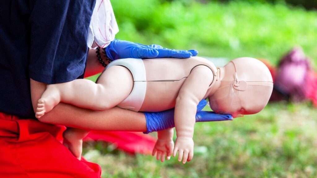 Person wearing blue gloves holds a baby CPR mannequin outdoors. The mannequin is positioned face-down, possibly demonstrating a rescue technique. The background is grassy, with some blurred red objects visible.