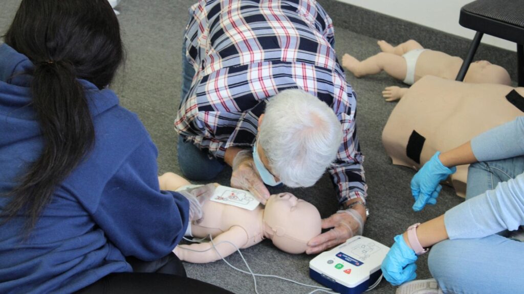 People are practicing CPR on infant mannequins on a carpeted floor. One person is placing an AED pad on a mannequin's chest while others observe, wearing gloves. A training AED device is visible on the floor.