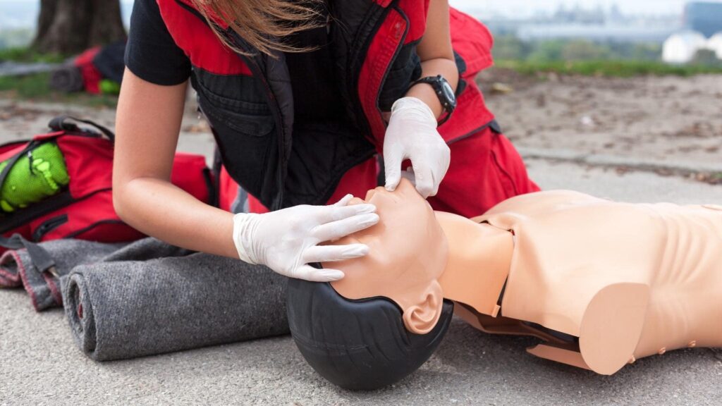 A person wearing gloves and a red vest practices CPR on a mannequin outdoors, with a red backpack nearby. The scene suggests a first aid training session on a concrete surface with blurred greenery in the background.