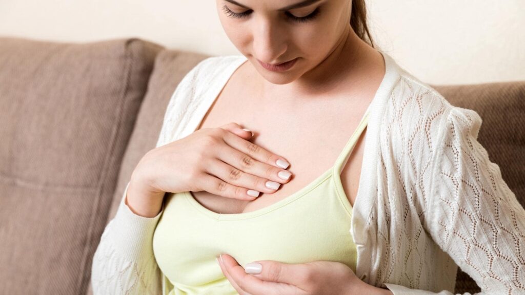 A woman wearing a light yellow top and white cardigan sits on a couch, looking down while gently touching her chest. She appears to be examining or massaging the area. Her fingernails are painted light pink.