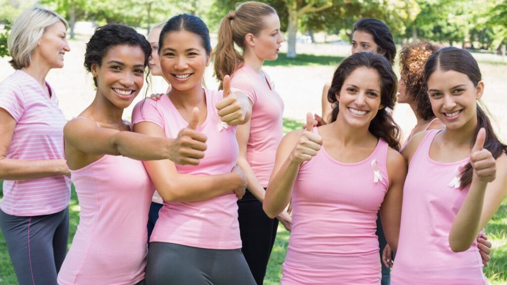 A group of women wearing pink tops and smiling, stand outdoors with trees in the background. They give thumbs up gestures, some with pink ribbons on their shirts, suggesting participation in a breast cancer awareness event.