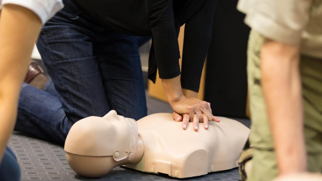 A person kneels on the floor, performing chest compressions on a CPR training mannequin. Another person is partially visible nearby, observing the practice.