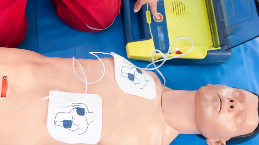 A person practices using an AED on a CPR training manikin. The manikin has electrode pads with illustrated placement instructions on its chest. A defibrillator is next to it, with a hand visible adjusting the device.