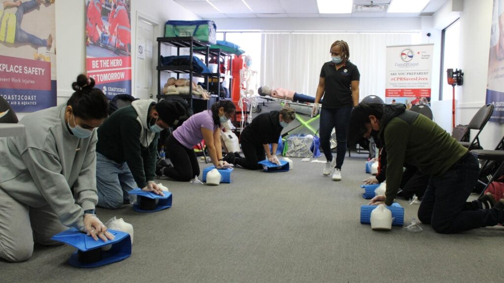 A group of people practice CPR on mannequins in a classroom setting. An instructor stands nearby observing. They are all wearing masks, and instructional posters are visible on the walls.