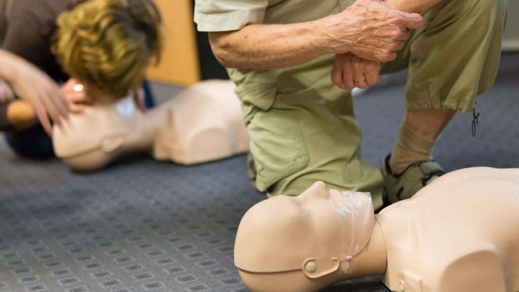 Two people practicing CPR on mannequins. One person is kneeling and compressing the chest of a mannequin, while the other prepares to administer rescue breaths. Both are focused on their training in a carpeted room.