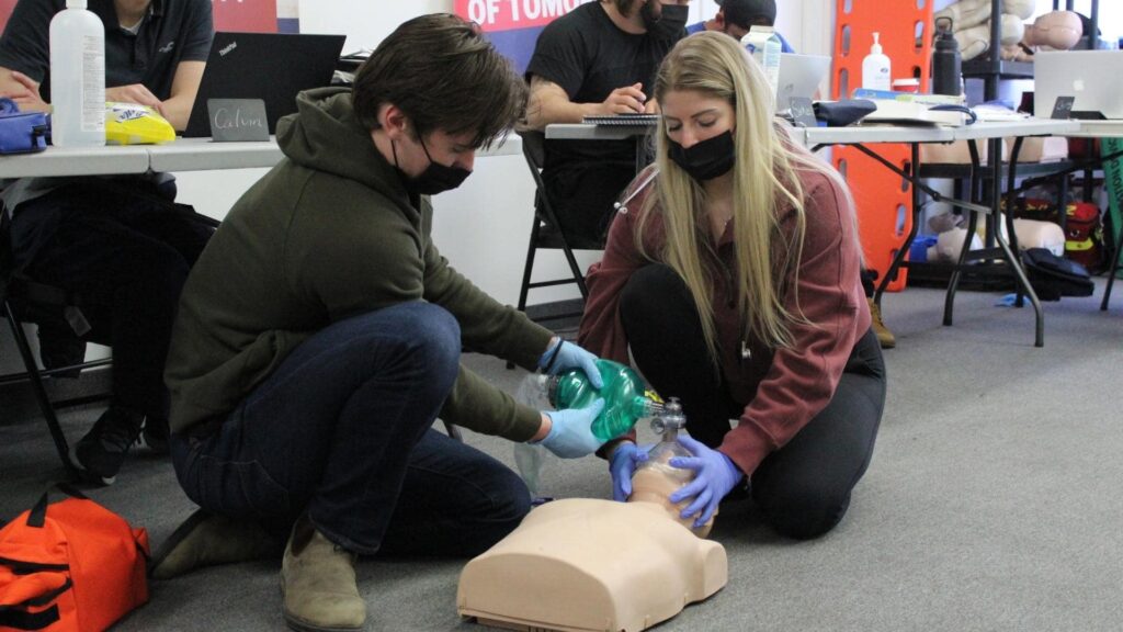 Two individuals in face masks and gloves are practicing CPR on a medical dummy. One is holding a bag valve mask over the dummy's face, while the other watches. Other participants and equipment are visible in the background.