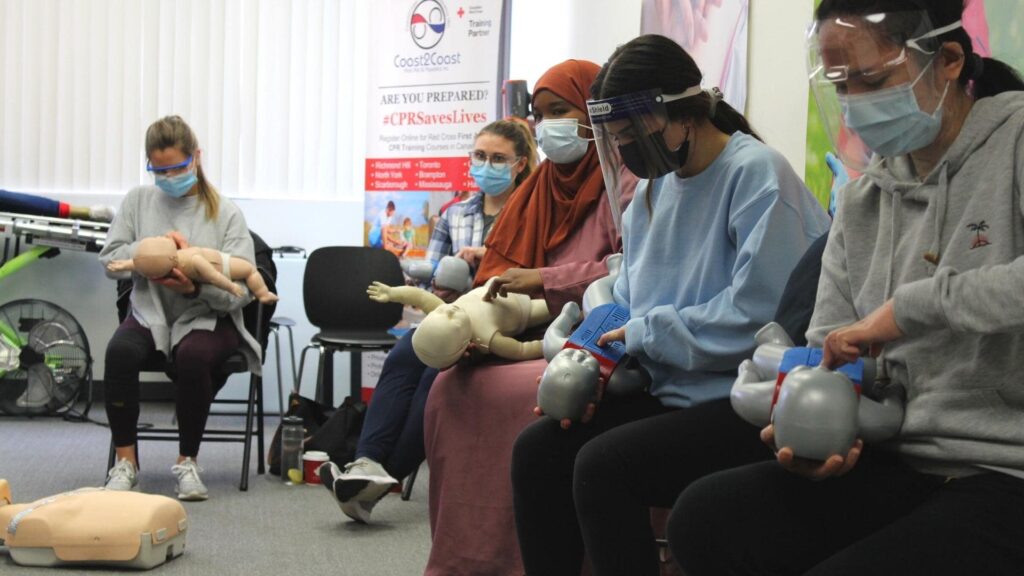 A group of people wearing masks and face shields are sitting in a classroom, practicing CPR techniques on infant mannequins. They are focused and engaged in the training session.