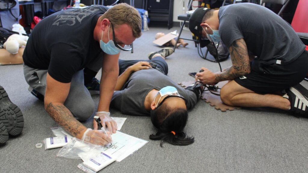 Two people wearing masks and gloves attend to a person lying on a carpeted floor. One person documents information while the other examines the patient. Various medical supplies are scattered nearby.