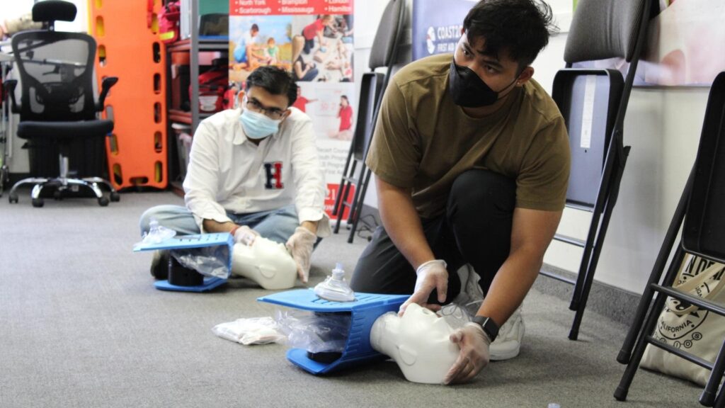 Two people wearing masks perform CPR on practice dummies in a classroom setting. They are kneeling on the floor with their hands positioned on the chests of the dummies. Instructional materials are visible in the background.