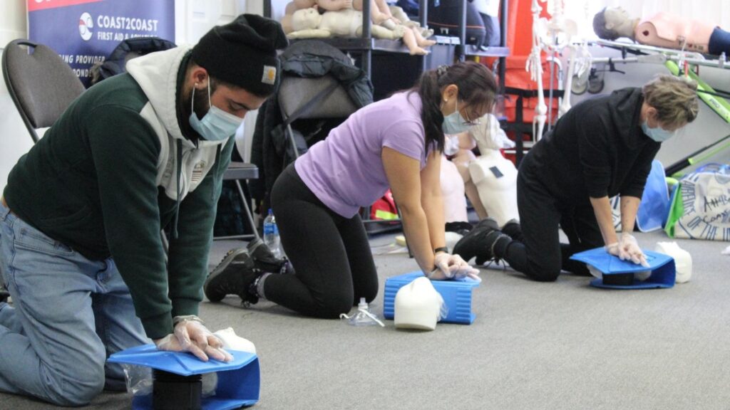 Three people wearing masks practice CPR on training mannequins in a classroom setting. They are kneeling and performing chest compressions. Medical equipment is visible in the background.