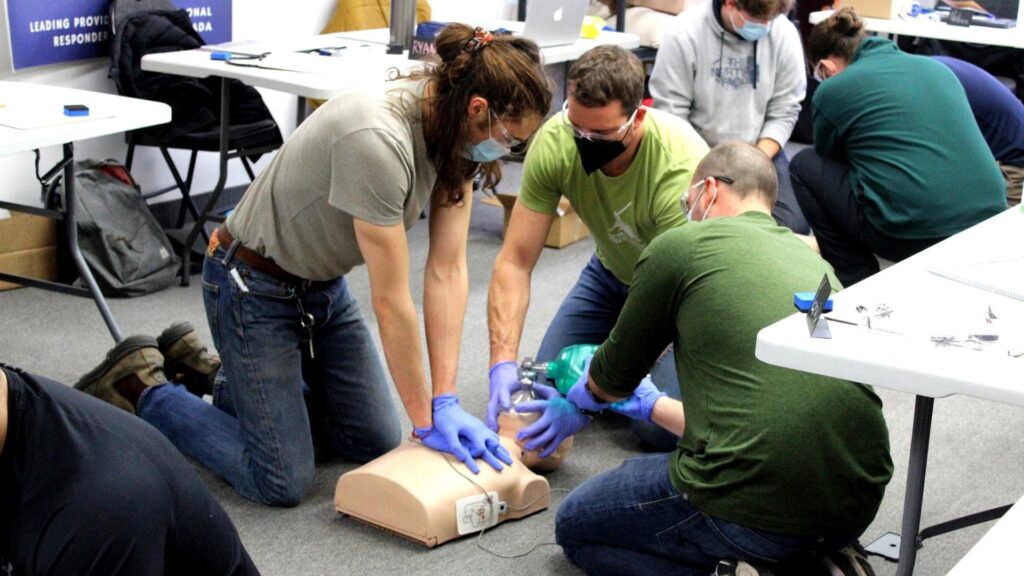 A group of people wearing masks are practicing CPR on a training mannequin. One person administers chest compressions while another uses a bag valve mask. Tables with equipment are in the background.