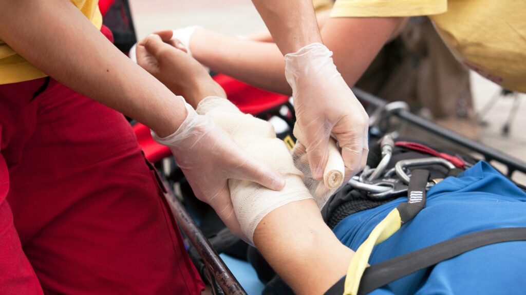 Medic wearing gloves bandaging a person's arm, who is wearing a blue garment and lying on a stretcher. Another person assists in the background. The scene suggests emergency medical care in progress.