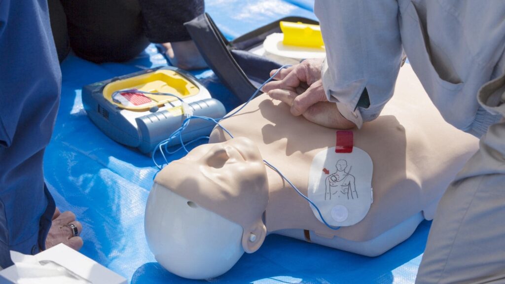 A person performs chest compressions on a CPR training mannequin. An automated external defibrillator (AED) with connected pads is visible beside the mannequin on a blue mat.