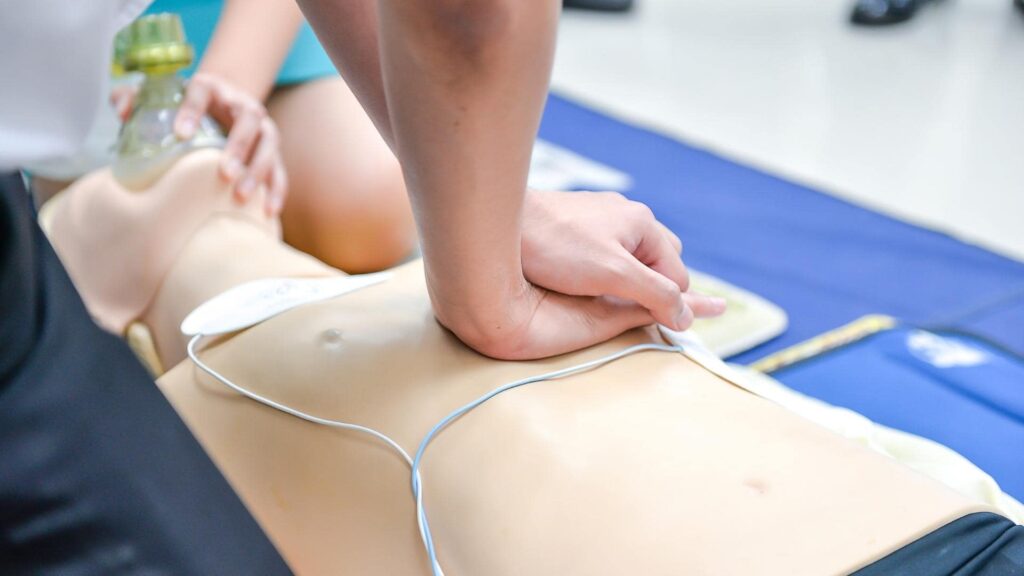 A person is performing chest compressions on a CPR manikin. Their hands are positioned on the chest, and the manikin is lying on a blue mat. A ventilation mask and other equipment are visible nearby.