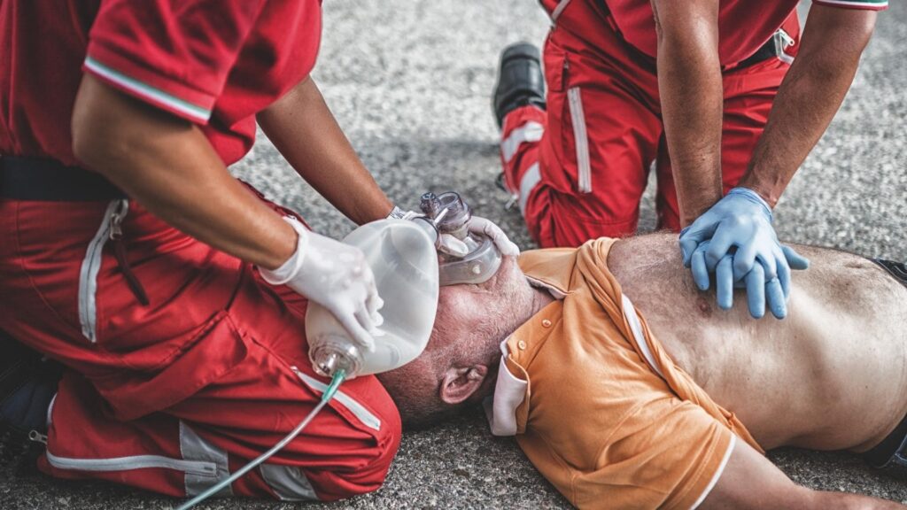 Two emergency responders in red uniforms perform CPR on a person lying on the ground. One administers chest compressions while the other holds an oxygen mask on the person's face. The person receiving aid wears an orange shirt.