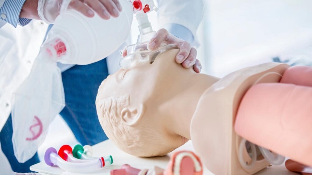 A medical professional demonstrates CPR on a realistic mannequin, using a bag valve mask for ventilation. Various airway management tools are visible in the foreground.