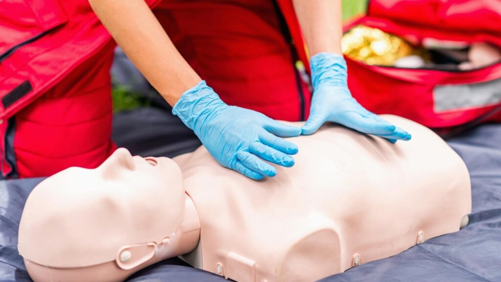 A person wearing blue gloves practices CPR on a medical mannequin. The mannequin is lying on a dark mat. A red emergency bag is partially visible in the background.