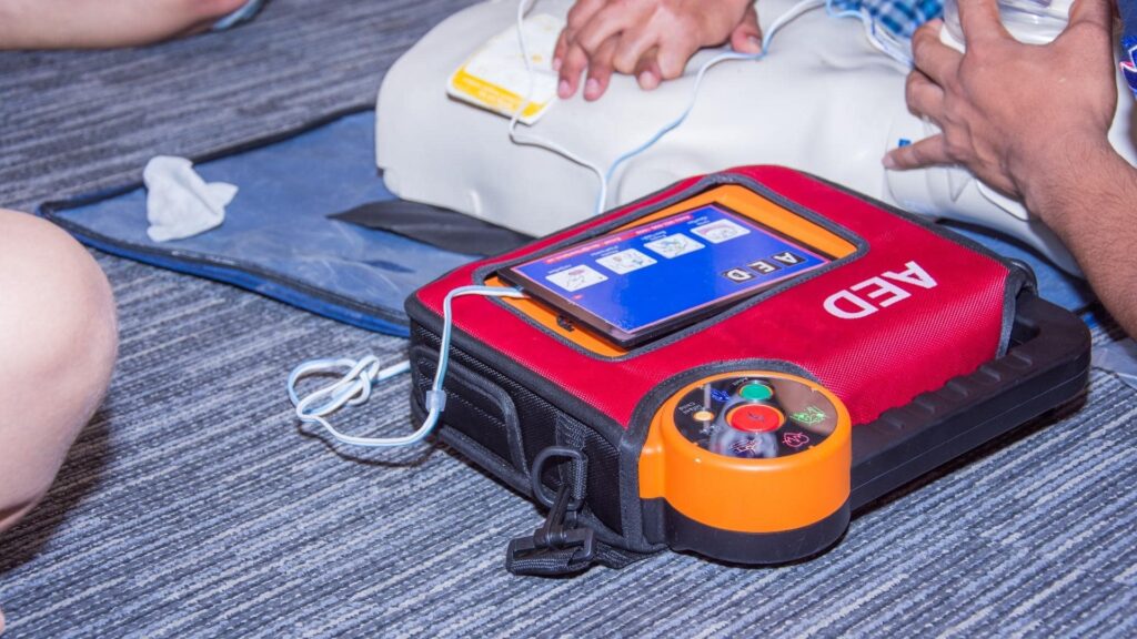 Two people practicing CPR with a training AED on a mannequin. One person adjusts the device while another's arm is visible. The AED displays instructions on a screen. The background shows a gray carpeted floor.