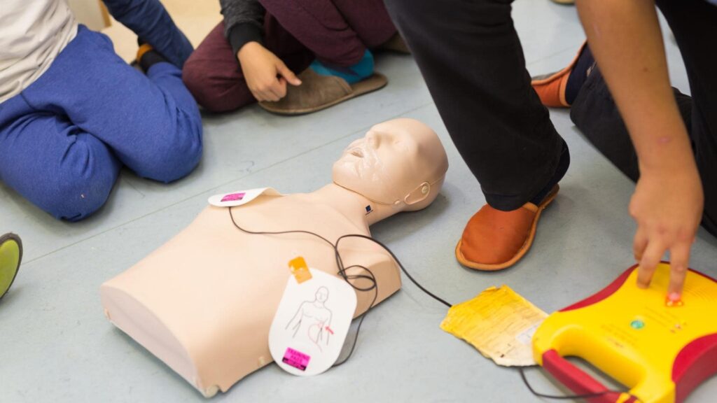 Children gather around a CPR training mannequin on the floor, with an automated external defibrillator (AED) placed nearby. The scene suggests a first aid or CPR training session.