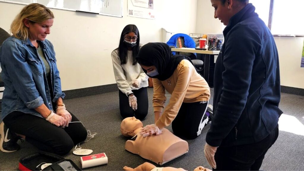 A group of four people practice CPR on a training mannequin in a classroom. One person compresses the mannequin's chest while others observe and assist. A defibrillator is placed nearby. They all wear gloves, and one person has a mask.