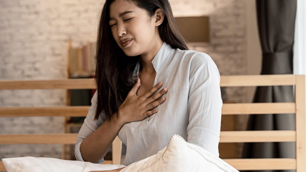 A woman sitting on a bed with her eyes closed and a pained expression, holding her hand to her chest. She is wearing a light gray blouse. The background includes a wooden headboard and a blurred lamp.