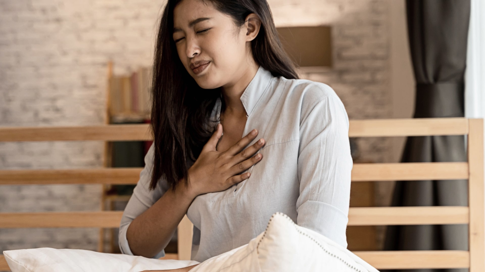 A woman sitting on a bed with her eyes closed and a pained expression, holding her hand to her chest. She is wearing a light gray blouse. The background includes a wooden headboard and a blurred lamp.