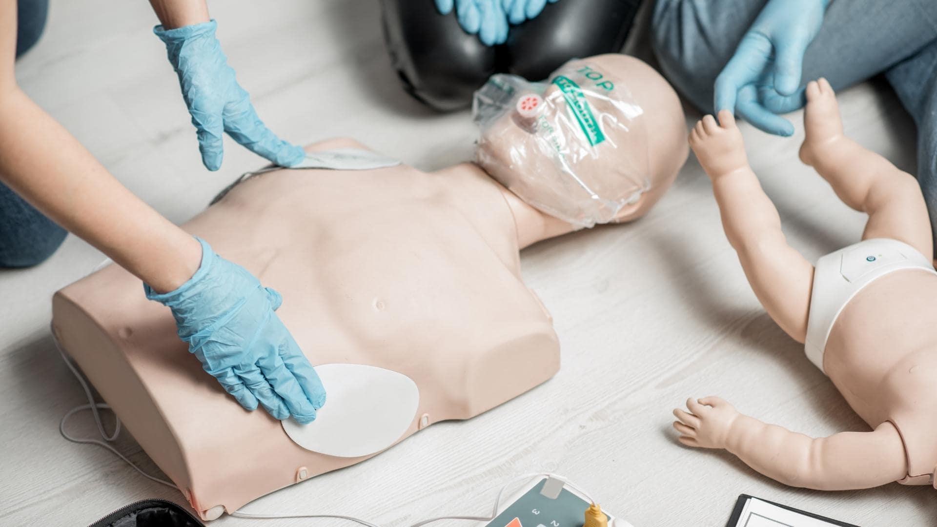 People wearing blue gloves practice CPR on adult and infant mannequins. The adult mannequin has defibrillator pads on its chest, while a defibrillator is visible nearby.