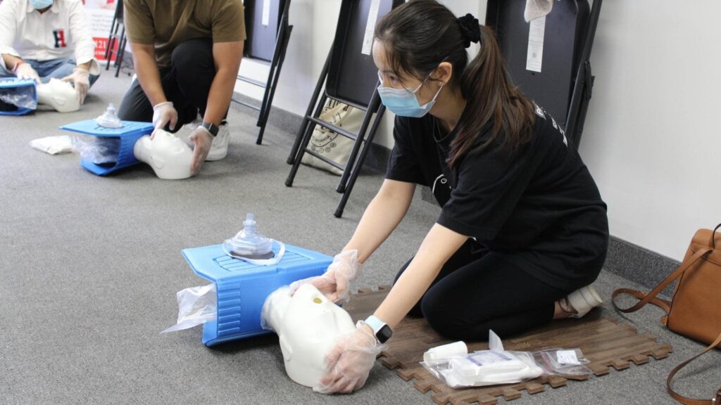 People in protective gloves and masks practice CPR on mannequins in a classroom setting. They are kneeling on the floor with medical supplies nearby. The focus is on a woman pressing down on a mannequin's chest.