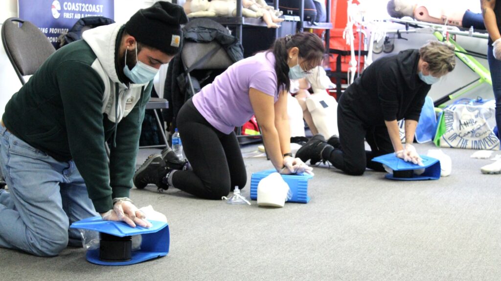 Three people kneel on the floor, practicing CPR techniques on blue training devices. They are wearing masks and gloves. A mannequin and first aid posters are visible in the background.