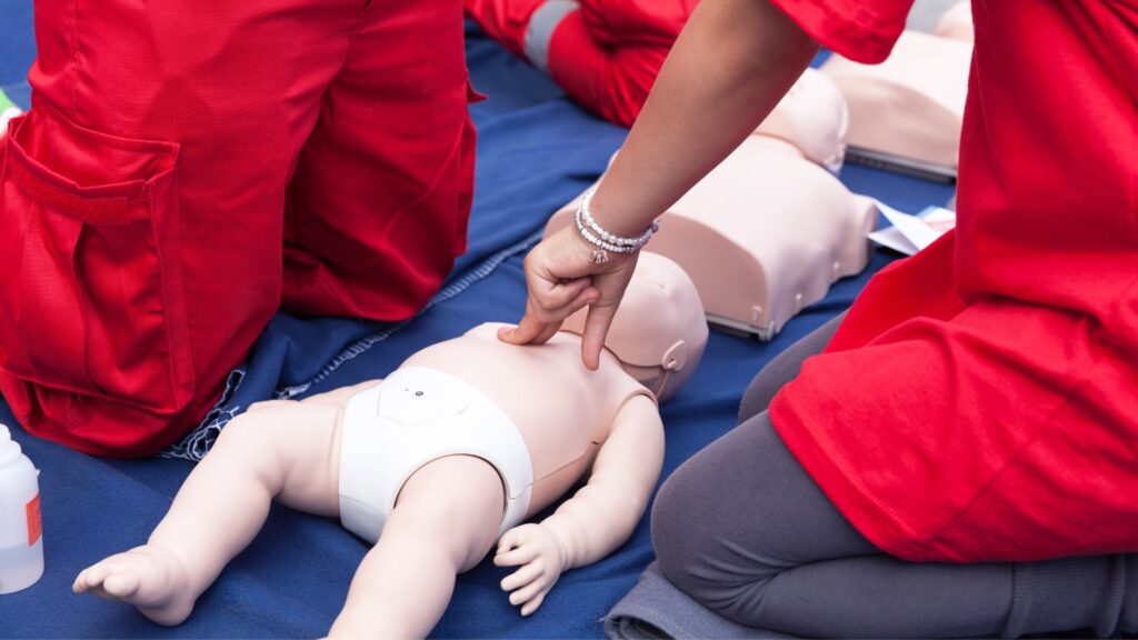 People in red shirts practice CPR on baby manikins placed on a blue mat. One person is giving chest compressions, focusing on proper technique during a first aid training session.