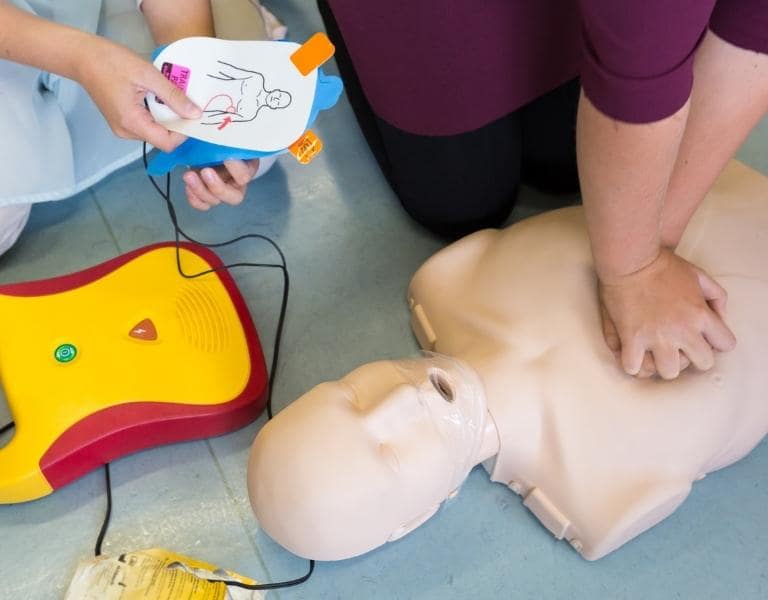 A person is practicing CPR on a mannequin, using both hands for chest compressions. Next to the mannequin, there is an AED with its pads being prepared for use. The scene takes place on a tiled floor.