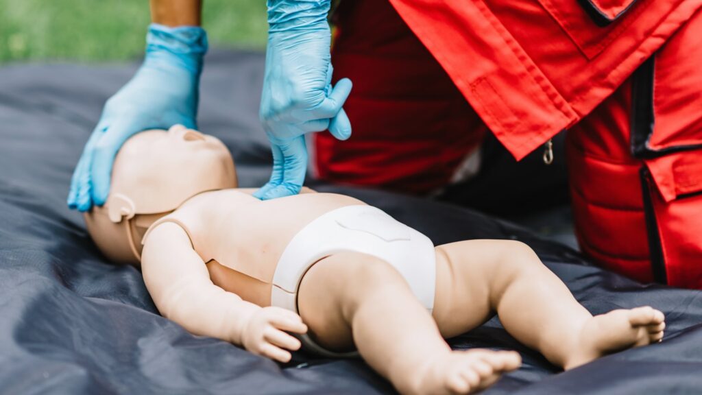 A person wearing blue gloves performs CPR on a baby mannequin dressed in a diaper. The mannequin is placed on a dark blanket, and the person is wearing a red uniform, suggesting a training or emergency scenario.