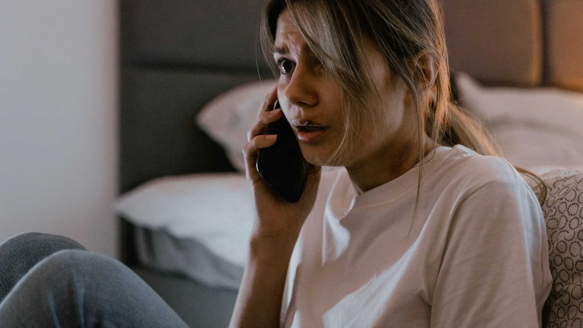 Woman sitting on a bed, holding a phone to her ear with a concerned expression. She has long hair and is wearing a white shirt and jeans. The background shows part of a bed with pillows. The lighting is warm and muted.