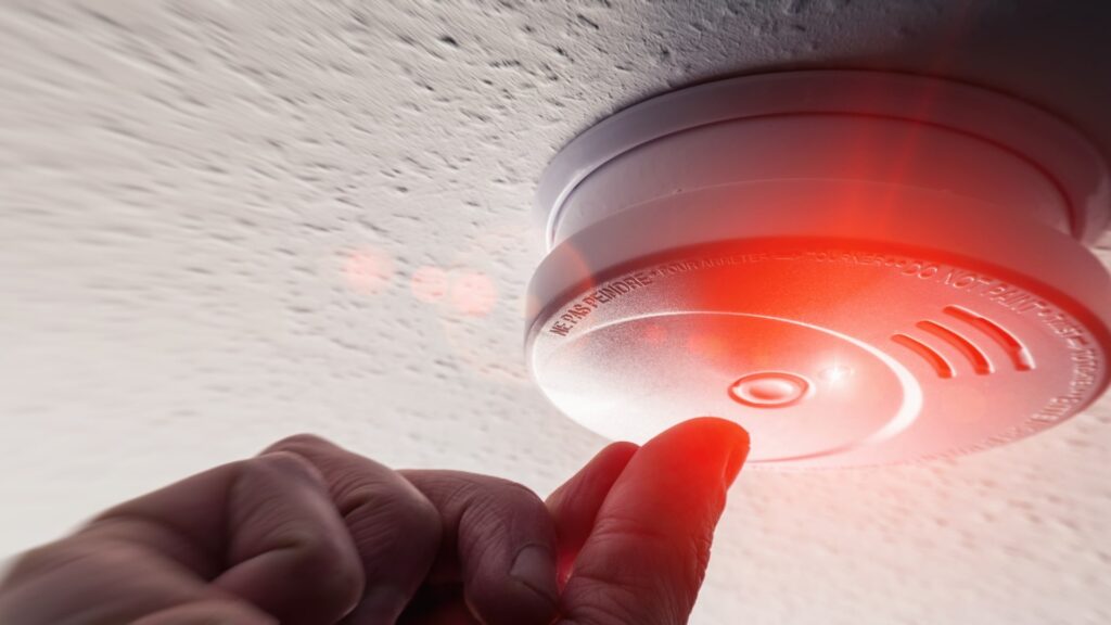 A hand reaches up to test a ceiling-mounted smoke detector. The device emits a red light, indicating active status. The ceiling surface has a textured finish.