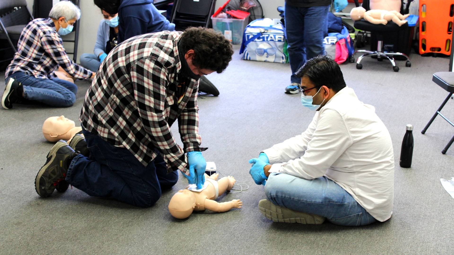People practicing CPR on infant mannequins. They're wearing masks and gloves in a classroom setting. Some are kneeling while others are seated, engaged in the activity, with training materials visible in the background.