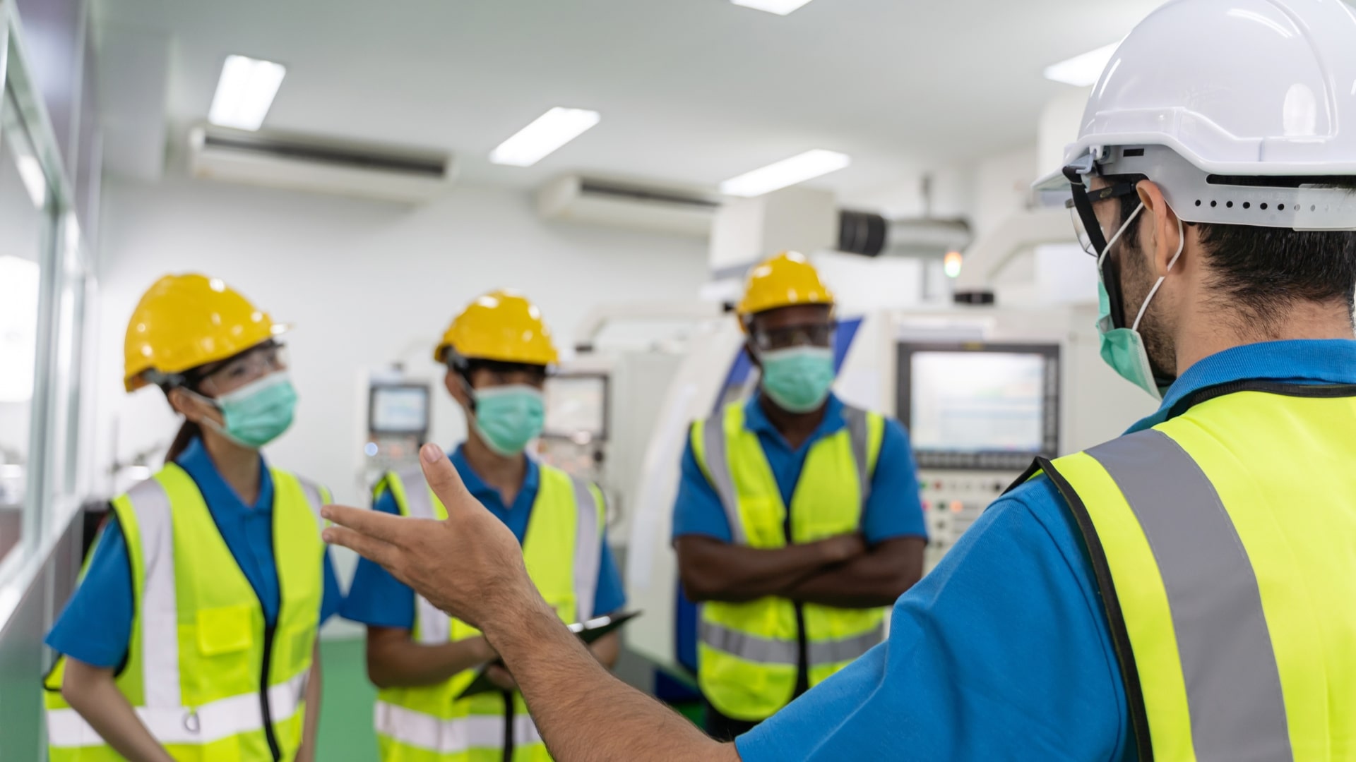 A person wearing a safety vest and helmet speaks to three others in similar attire. All are wearing face masks and standing in an industrial setting with machinery in the background.