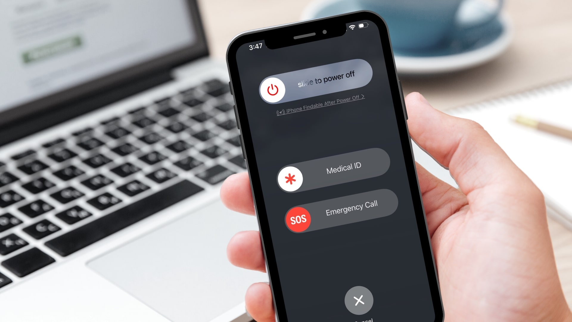 A person holds an iPhone displaying the "slide to power off" screen with options for "Medical ID" and "Emergency Call." A laptop keyboard and notebook are blurred in the background.