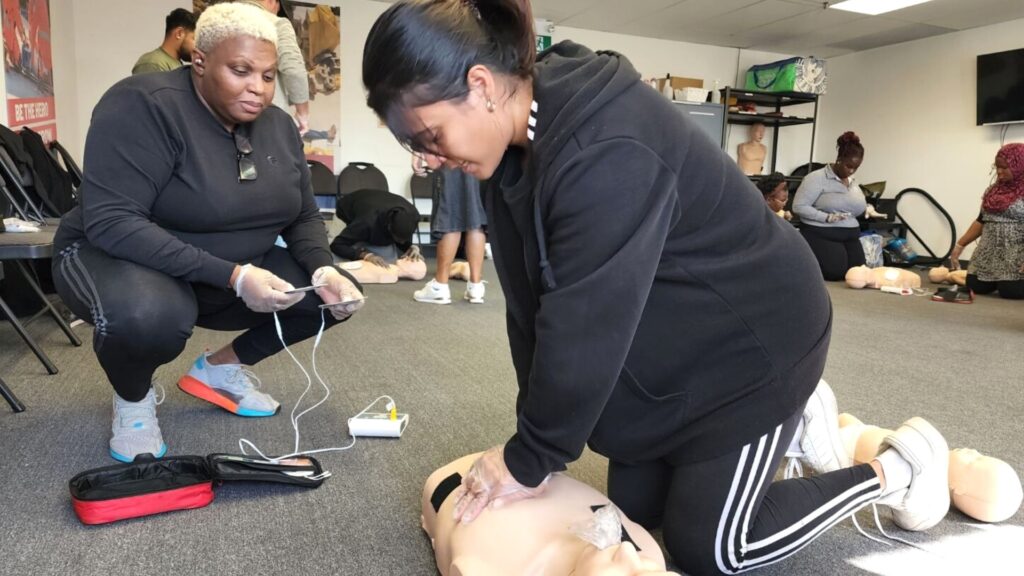 A person performs CPR on a dummy while another observes, holding a medical device. Several other CPR dummies and participants are visible in the background of the training room.