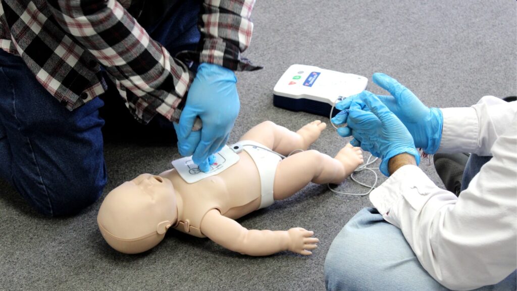 Two individuals wearing blue gloves are practicing CPR on an infant training mannequin. One person is placing defibrillator pads on the mannequin's chest while the other prepares an AED device in a room with a gray carpet.