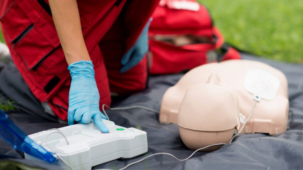 A person wearing red medical attire and blue gloves is kneeling on a mat outdoors next to a CPR dummy. They are pressing a button on a defibrillator device, preparing for resuscitation practice. A red bag is in the background.