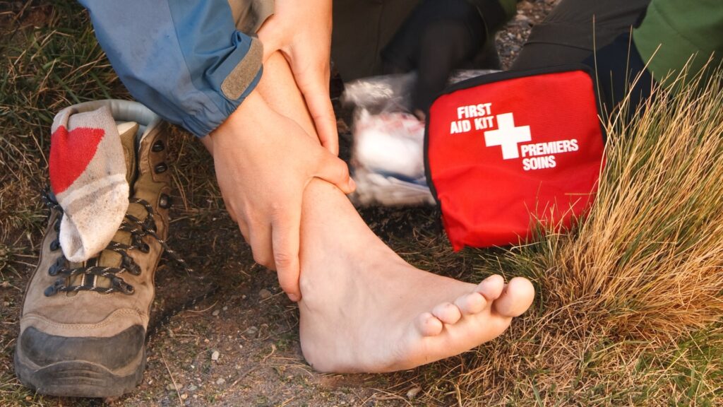 A person is sitting on the ground with their bare foot exposed, holding their injured ankle. A hiking boot lies nearby. A red first aid kit is open beside them, indicating they are tending to the injury outdoors.