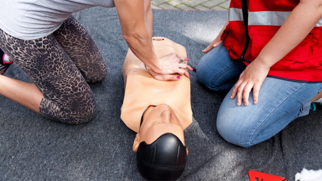 Two people perform CPR on a mannequin. One person presses on the chest while the other observes. Both are kneeling on a gray mat, with the second person wearing a red vest. The mannequin is tan with a black head.